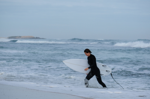Female surfer in wetsuit walking out of ocean holding shortboard surfboard with coastal background - Australian Stock Image