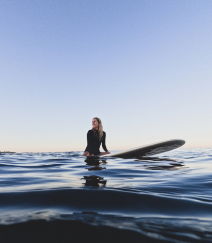 Female surfer sitting on longboard surfboard in springsuit with glassy blue ocean and clear sky - Australian Stock Image