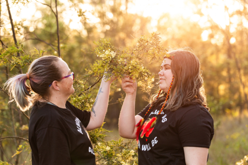 First Nations Australian mother and teen daughter with wattle flowers in bushland - Australian Stock Image