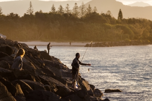 Fisherman standing on rocks beside a river at sunset - Australian Stock Image