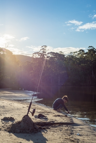 Fishing rod and boy playing in river outlet with sun flare trees and blue sky - Australian Stock Image