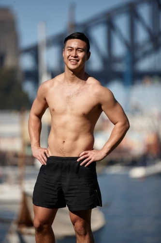Fit Asian man smiling with hands on hips in front of harbour bridge - Australian Stock Image
