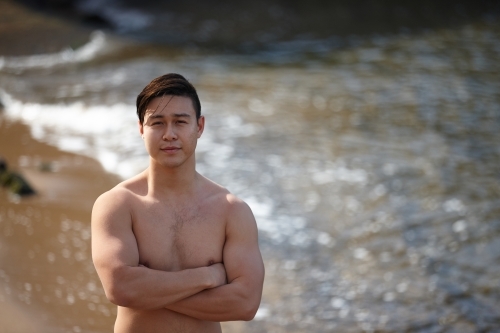 Fit Asian man with arms folded at beach - Australian Stock Image