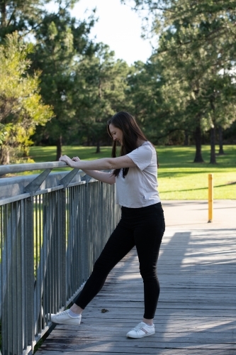 Fit young woman in casual clothes exercising at railing on foot bridge in park. - Australian Stock Image