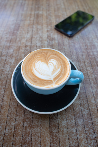 flat white coffee in a wide blue coffee cup with black saucer, with a phone in the background - Australian Stock Image