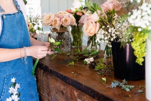 Florist preparing wedding buttonholes and small corsages at a work bench full of flowers - Australian Stock Image