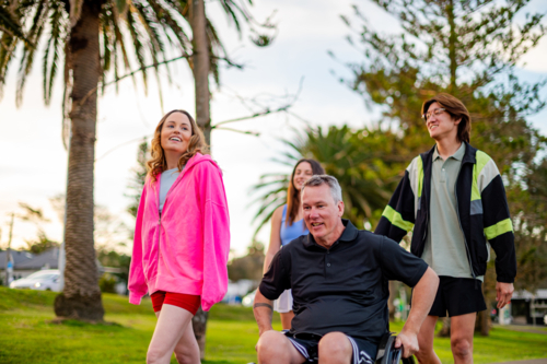 Four friends casually walking in a park near palm trees at sunset, laughing and chatting together - Australian Stock Image