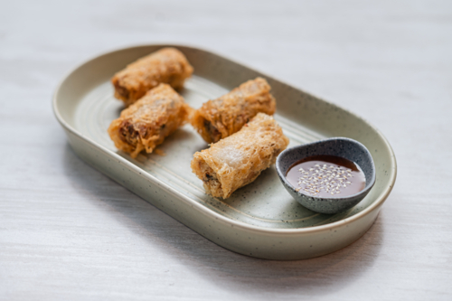 Four golden crispy spring rolls on plate alongside a small bowl of sauce - Australian Stock Image