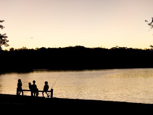 Four young children silhouetted against water with fishing rod - Australian Stock Image