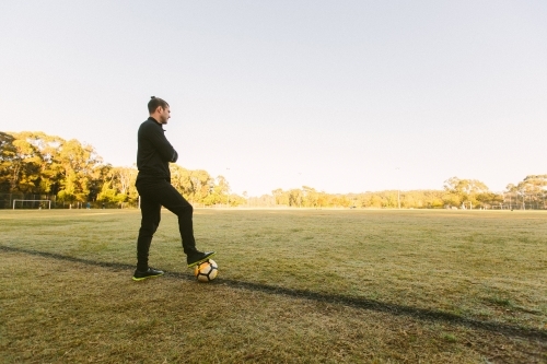 Full body shot of a young man standing on a big field stepping on a soccer ball with one foot - Australian Stock Image