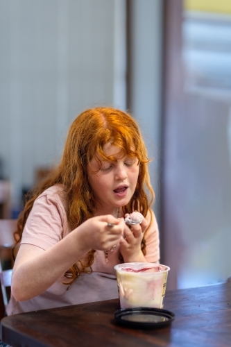 ginger-haired girl eating flavoured yoghurt on a spoon from plastic tub - Australian Stock Image