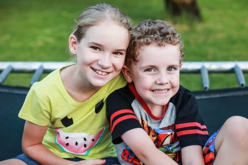 Girl and boy sitting close together on trampoline smiling - Australian Stock Image