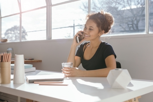 Girl in her 20s talking on a mobile phone at a desk in a light bright office - Australian Stock Image