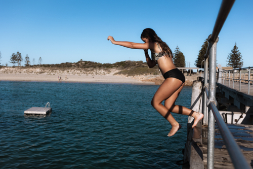 Girl jumping from jetty into the ocean bathed in golden summer sun - Australian Stock Image