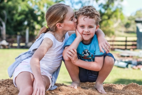 Girl kissing boy on forehead sitting on pile of dirt - Australian Stock Image