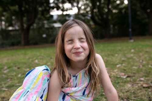 Girl pulling silly face with eyes closed sitting outside on grass - Australian Stock Image