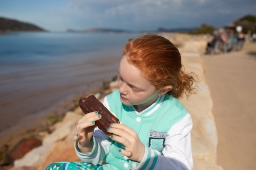 Girl sitting on a sandstone pathway eating a chocolate eclair - Australian Stock Image