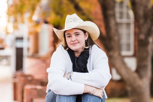 Girl sitting on brick wall hugging knees wearing hat smiling - Australian Stock Image