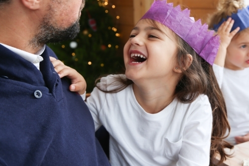 Girl sitting on dad's lap laughing wearing paper hat - Australian Stock Image