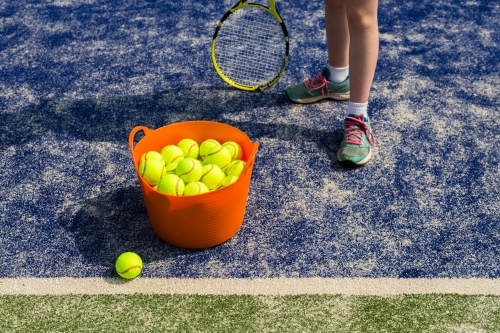 Girl standing next to an orange basket of tennis balls on a blue tennis court - Australian Stock Image