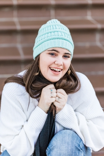 Girl wearing beanie clasping scarf smiling sitting on steps - Australian Stock Image