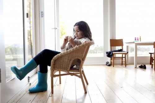 Girl wearing blue gumboots sitting inside on a wicker chair - Australian Stock Image