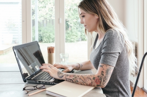 Girl with tattoos typing on computer keyboard at home - Australian Stock Image