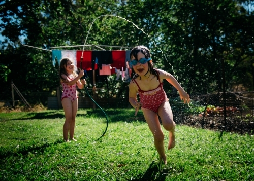Girls playing under hose, brunette girl running under water with goggles on - Australian Stock Image