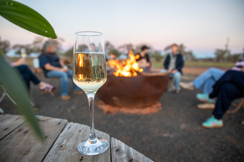 Glass of sparkling wine on a wooden table, with a crowd of women around a firepit in the background - Australian Stock Image