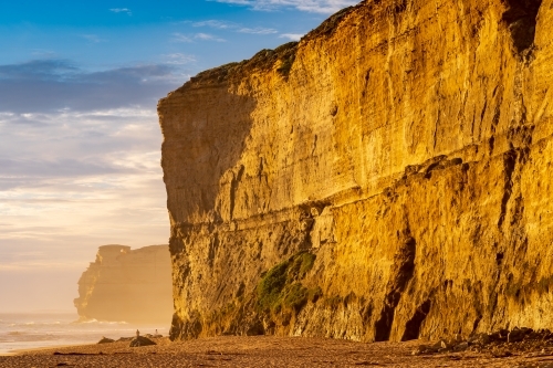 Golden light on the face of a vertical cliff face rising from a sandy beach - Australian Stock Image