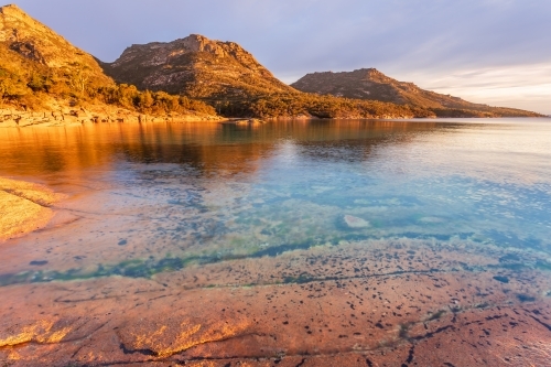 Golden lighting on coastal mountains behind calm water in large rock pools - Australian Stock Image