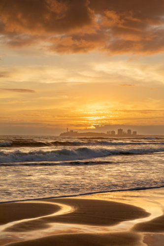 Golden sunrise at the beach with sand reflections, waves and buildings in background - Australian Stock Image