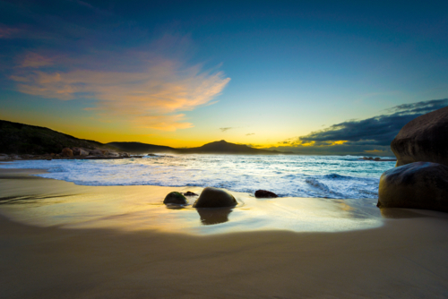 Golden sunset light reflecting off wet sand at a secluded beach with gentle waves and rocks - Australian Stock Image