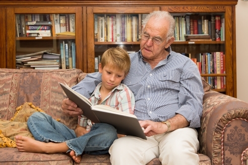 Grandfather and grandson reading a book on a sofa together - Australian Stock Image