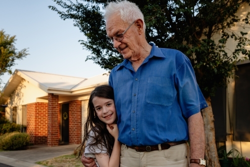 Grandfather hugs his granddaughter adoringly in front of his house