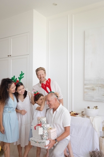 Grandson putting reindeer antlers on grandfather at Christmas, with family looking on - Australian Stock Image