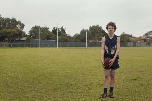 Grassroots Footy player standing with ball on local football ground - Australian Stock Image