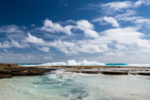 Ground level view of water flowing into rockpool with waves behind and dramatic blue cloudy sky