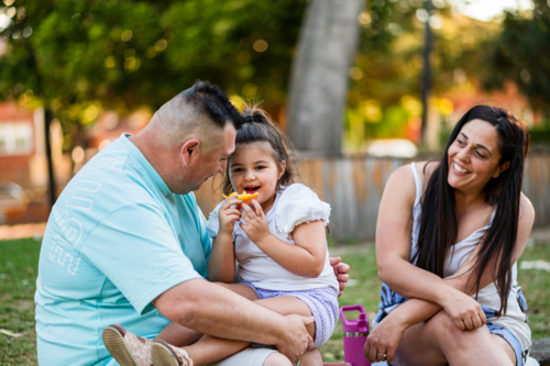 Group of three people share food and laughter while sitting on grass in a park - Australian Stock Image