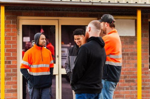 Group of workmen mechanics chatting together outside office at workplace some in high-vis - Australian Stock Image
