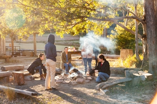 Group of young people relaxing around campfire in bushland on sunny winter morning - Australian Stock Image