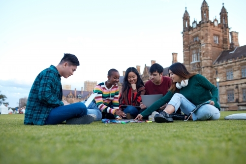 Group of young university students hanging out sitting on grass studying and using devices