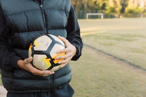 Half body shot of a man wearing black vest holding a soccer ball with two hand, standing on a field - Australian Stock Image