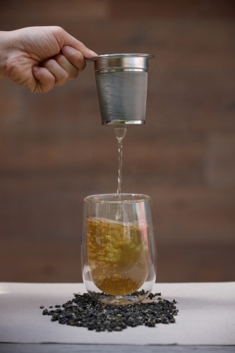 Hand pouring hot tea through strainer into glass cup - Australian Stock Image