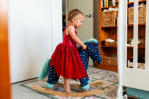 Happy Aussie kid in red dress riding on rocking horse toy in her bedroom - Australian Stock Image