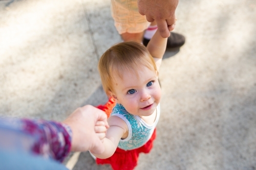 Happy baby girl holding mum and dad's hands walking outside - Australian Stock Image