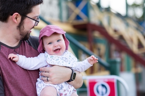 Happy baby wearing hat outside in dads arms with grandstand behind at showground - Australian Stock Image