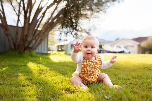 Happy baby with hand up giving high five outside on grass - Australian Stock Image