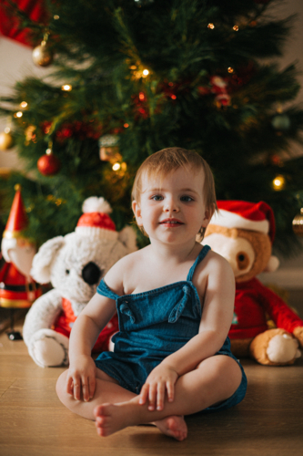 happy blonde toddler smiles by a teddy bear and Christmas tree - Australian Stock Image
