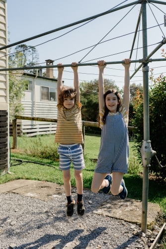 Happy brother & sister hanging from a Hills Hoist clothes line in an Australian country backyard - Australian Stock Image